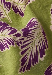 Tessuto di cotone con uno sfondo verde e grandi motivi di foglie viola e bianche. La texture appare morbida e leggermente stropicciata.