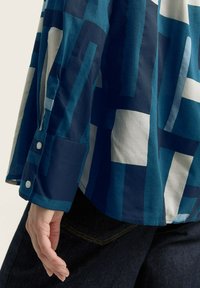 Camicia a maniche lunghe blu e bianca con un polsino abbottonato, caratterizzata da forme geometriche e colori a contrasto, realizzata in un tessuto morbido e leggero.