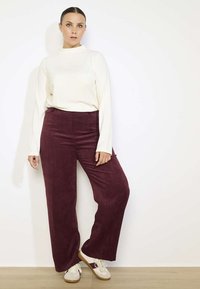 Top a maniche lunghe crema abbinato a pantaloni a gamba larga in velluto a coste bordeaux. L'outfit è completato da sneakers bianche su una superficie in legno chiaro.