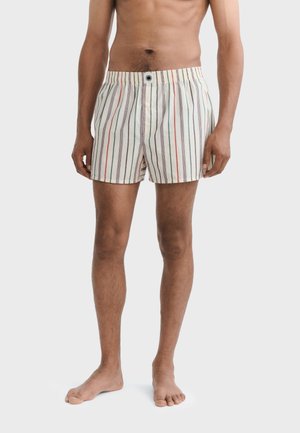 Mann trägt gestreifte Boxershorts in Creme mit roten, grünen und grauen vertikalen Linien, steht barfuß vor einem einfarbigen Hintergrund.