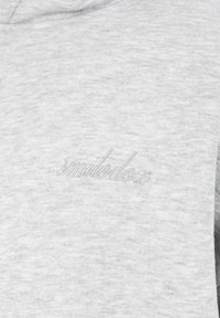 Lichtgrijze stof met subtiel geborduurde cursieve tekst "Imelodoci" in bijpassende draad op een zacht, gestructureerd kledingstuk.