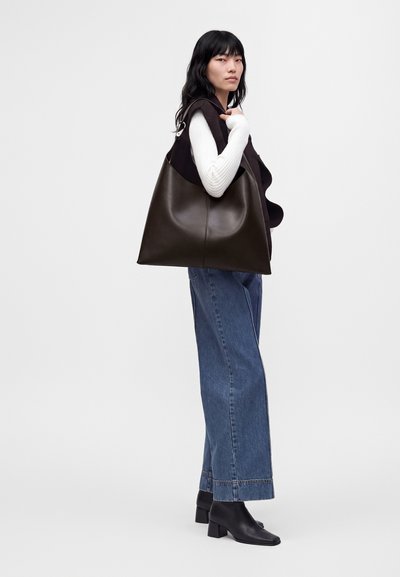 Femme portant un pull blanc, un gilet noir, un jean bleu et des bottes noires, portant un grand sac fourre-tout en cuir marron foncé sur l’épaule.