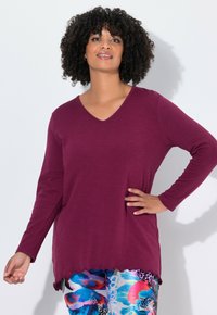 Burgunderfarbene Langarmbluse mit V-Ausschnitt und gewelltem Saum, hergestellt aus einem strukturierten Stoff, kombiniert mit gemusterten, bunten Leggings.