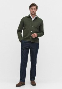 Grön ullblandad jacka med ett litet bröstfick, knäppning fram, bärs över en vit skjorta, i kombination med marinblå byxor och bruna skor.