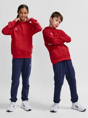 Dos niños con sudaderas rojas a juego, pantalones azul marino y zapatillas blancas sobre un fondo gris liso.
