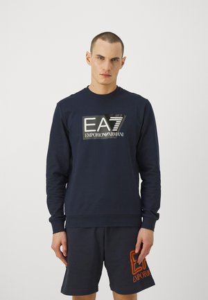 Felpa blu navy con colletto rotondo, maniche lunghe e un design di logo metallico. Abbinata a pantaloni corti scuri coordinati con un logo arancione.
