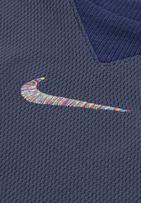 Temno modra polosrajca iz teksturirane tkanine z V-izrezom in barvitim vezenim logotipom Nike, ki ima vrtinčast vzorec.