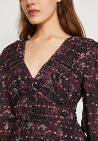 Vestido floral escuro com um decote V profundo, mangas bufantes e fecho com botão. Apresenta tons variados de roxo, detalhes em dourado e textura plissada.