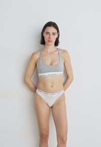 Grijze katoenen sportbeha met een logo band en gestreepte witte stringonderbroek met een logo tailleband. Model staat met het gezicht naar voren.