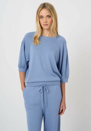 Blondynka w jasno niebieskim dzianinowym topie z rękawami do łokci i pasujących spodniach ze ściągaczem, stojąca na jednolitym tle.