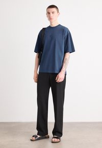 Camisa de manga corta azul marino con acentos de tela plisada, combinada con pantalones negros y sandalias negras con una correa de hebilla.