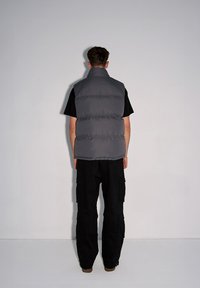 Szary kamizelka z podpinką o wysokim kołnierzu, noszona na czarnej koszuli. Ma gładką teksturę i puchate sekcje, w połączeniu z czarnymi spodniami cargo.