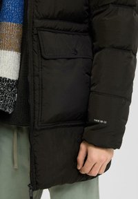 Čierna puffová bunda so zipsom vpredu, bočnými vreckami a rebrovanými manžetami; na rukáve je nápis "TOTO JE QS" bielou farbou; doplnená viacfarebným šálom.