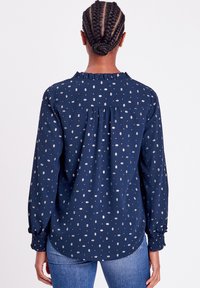 Marinebluse mit Rüschensaumkragen, langen Ärmeln und geknöpften Manschetten. Verfügt über ein weißes geometrisches Muster.