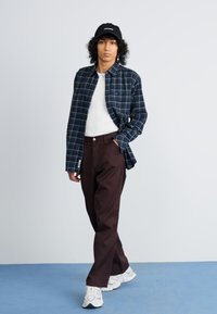 Plaid flannel shirt in navy and green, white T-shirt, brown wide-leg pants, and white sneakers. Curly hair, black cap with text.