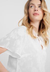 Blusa blanca con bordado floral, mangas cortas y detalle de lazo en el cuello. Tejido ligero y texturizado con un aspecto translúcido.