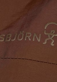 Brun stof med præget tekst "SBJÖRN" sammen med et simpelt, stiliseret figurikon. Glat tekstur med synlige syninger.