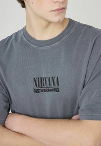 T-shirt en coton gris arborant le texte imprimé "NIRVANA NEVERMIND" en noir. Encolure ronde et manches courtes, avec une texture douce.
