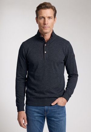 Man met kort bruin haar, gekleed in een zwarte pullover en blauwe spijkerbroek, staand tegen een effen lichte achtergrond.