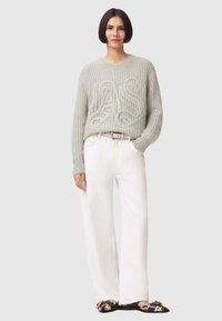 Hellgrauer Strickpullover mit einer strukturierten Vorderseite, kombiniert mit weißen, weit geschnittenen Hosen und gemusterten Schuhen mit dunklen Akzenten.