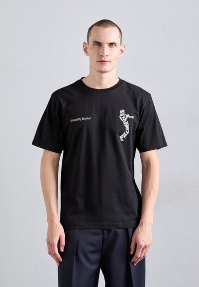 Svart bomull t-shirt med korta ärmar. Har texten "United By Rhythm" och en design av en dansare på framsidan. Enfärgad baksida.