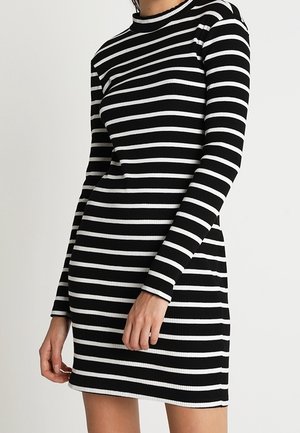 Femme portant une robe noire ajustée à manches longues avec des rayures blanches horizontales, debout devant un fond clair uni.