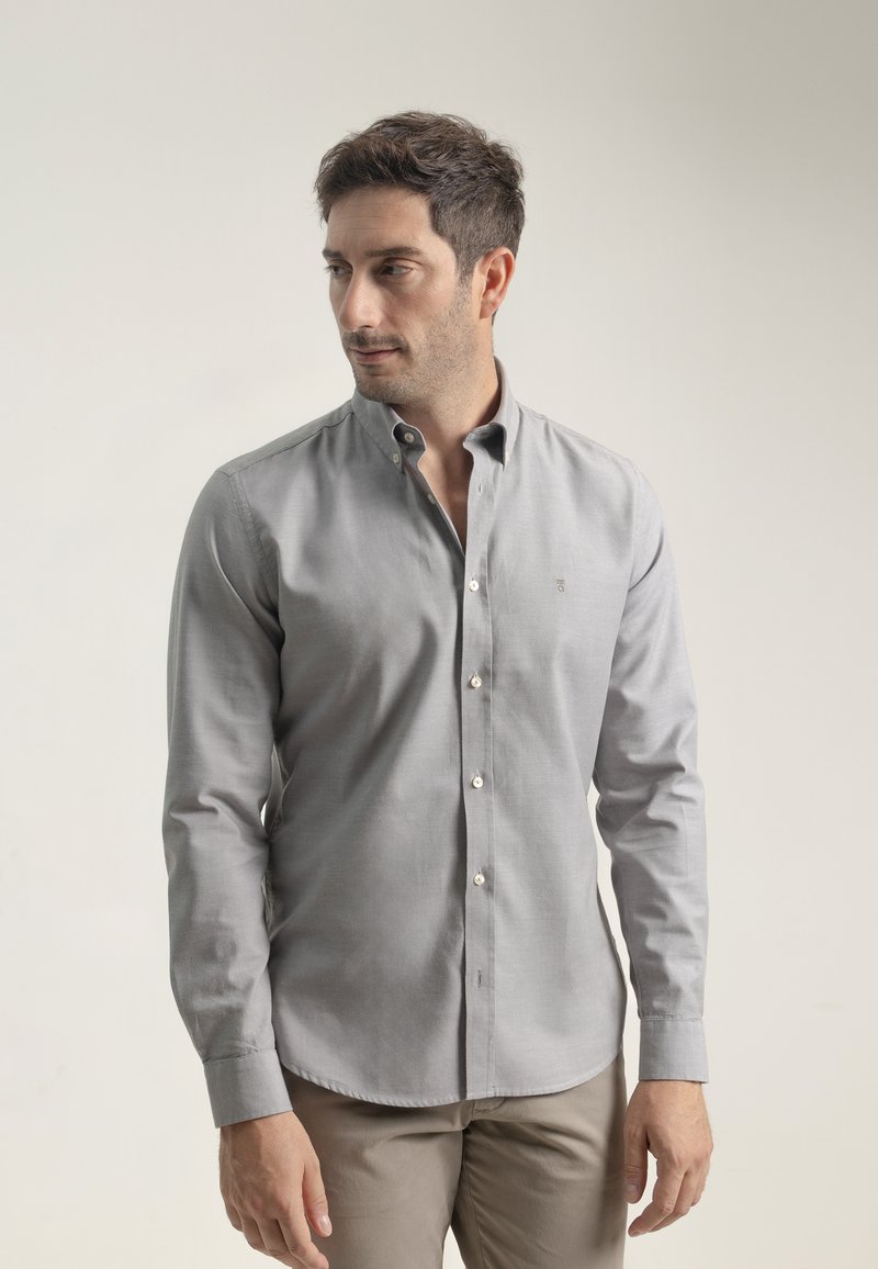 Camisa gris de botones con cuello, que presenta una textura sutil y un pequeño logo en el pecho. Mangas largas con puños abotonados.