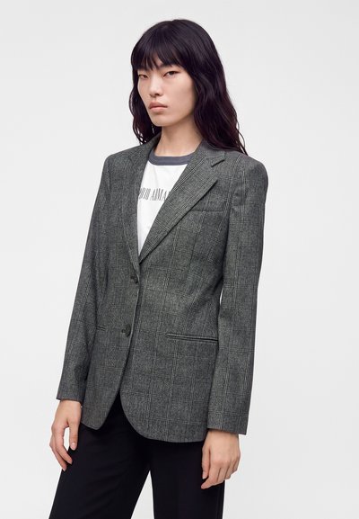 Femme aux longs cheveux foncés portant un blazer à carreaux gris sur un t-shirt blanc graphique et un pantalon noir, sur un fond clair uni.