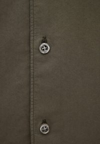 Zbliżenie na dwa szare guziki przymocowane do oliwkowozielonej tkaniny z widocznym przeszyciem wzdłuż szwu przy guziku.