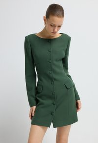 Eine Frau trägt ein tailliertes, langärmeliges, dunkelgrünes Blazer-Kleid mit Knöpfen und Klapptaschen, während sie vor einem schlichten, hellen Hintergrund steht.