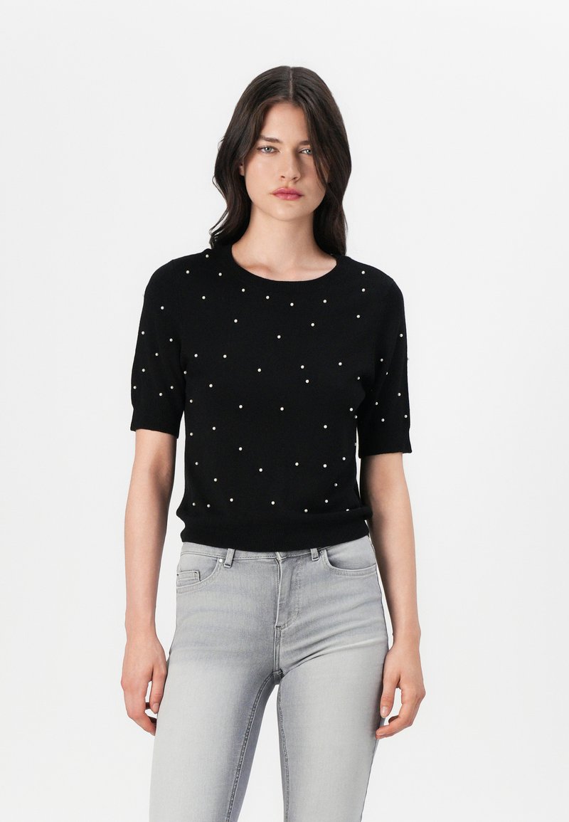 Pull en tricot noir à manches courtes avec un col rond et des perles blanches éparpillées en accent. Associé à un jean gris clair ajusté.