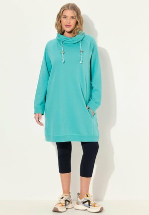 Vestido tunic de cuello de cowl en color turquesa, con mangas largas, bolsillos laterales y cordones texturizados. Combinado con leggings capri en azul marino y zapatillas deportivas.