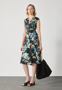 Robe noire à motifs floraux avec un design croisé, des manches courtes et des motifs floraux colorés. Associée à un sac en suède noir et des sandales à talons noires.