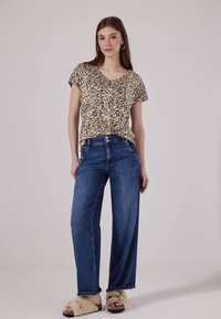 Kurzärmliges Oberteil mit Leopardenmuster, lockere Passform, kombiniert mit weiten blauen Jeans und flauschigen Slide-Sandalen. Heller Hintergrund.