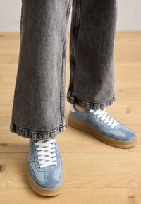 Heleda sinine nahast sneakers valgete paelte ja kummist tallaga, koos hallide avara säärega teksadega, millel on kulunud serv ja nähtavad õmblused.
