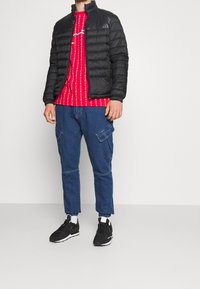 Chaqueta acolchada negra, camiseta roja con patrón blanco, pantalones cargo azules y zapatillas negras. Equipado con puños elásticos y bolsillos.