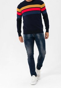 Pull marine à rayures horizontales jaunes, rouges et bleues. Associé à un jean foncé et des baskets blanches. Design décontracté, texture tricotée.