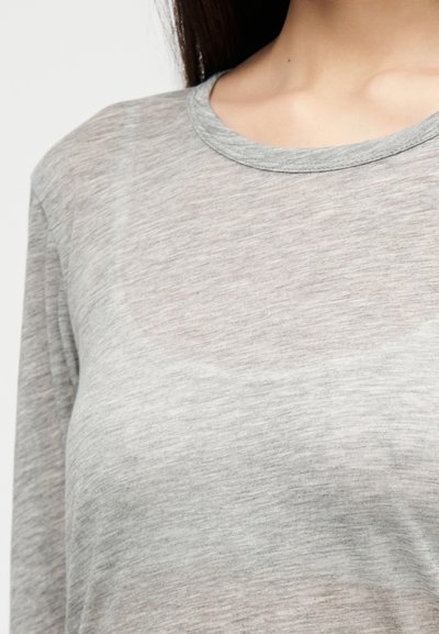 Detailný záber osoby nosiacej svetlosivú tričko s dlhými rukávmi a okrúhlym výstrihom, zobrazujúci rameno a hornú časť hrudníka.