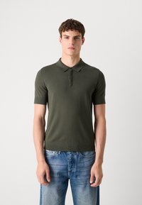 Olivengrønn t-skjorte med korte ermer i ribbestrikket tekstur, krage og tre-knapps knappestolpe, kombinert med lyseblå denimjeans.