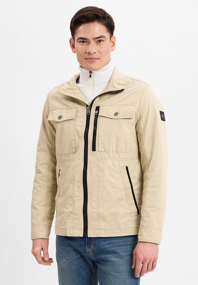 Beige leichte Jacke mit Reißverschluss vorne, zwei Brusttaschen, Seitentaschen und schwarzen Akzenten. Glattes Material und lässiger Schnitt.
