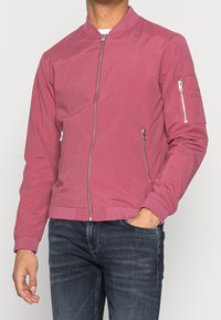 Muž v růžovém bomberu s kapsami se zipem na přední straně a rukávech, v kombinaci s tmavě modrými džíny, ruka v kapse.