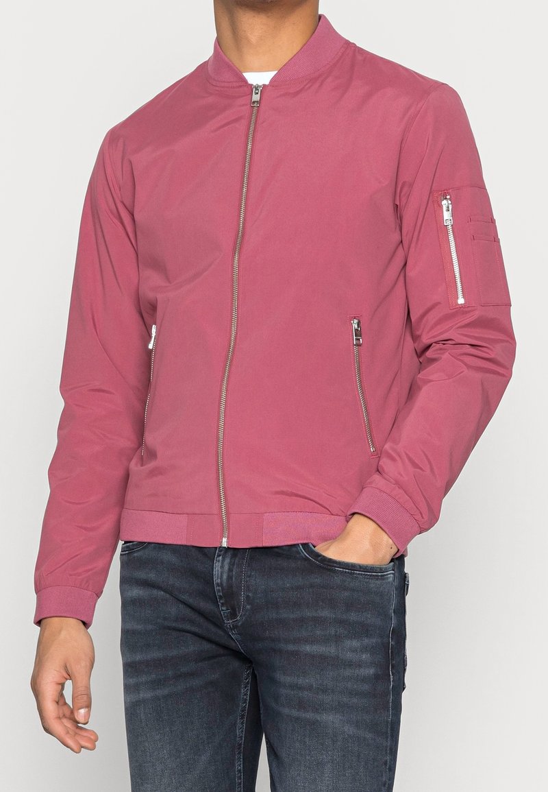Muž v růžovém bomberu s kapsami se zipem na přední straně a rukávech, v kombinaci s tmavě modrými džíny, ruka v kapse.