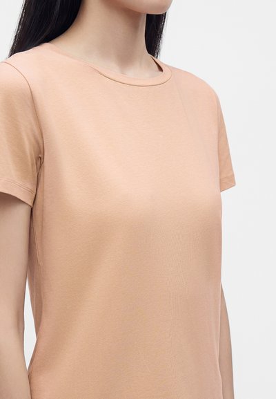 Femme aux longs cheveux foncés portant un t-shirt uni couleur pêche à manches courtes et col rond sur fond blanc.