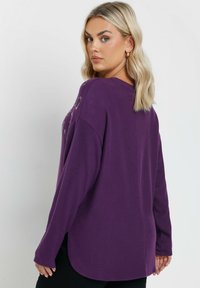 Übergroßes lila Oberteil mit langen Ärmeln, rundem Halsausschnitt und seitlichen Schlitzen, versehen mit einer weichen Textur und einem etwas längeren Rückensaum.