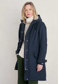Marinel blauw lange jas met een capuchon, voorzien van een ritsluiting, drukknoopsluiting en voorzakken. Voering met een zachte, textuurrijke binnenvoering.