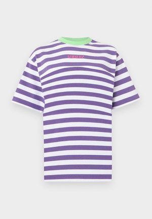 Osoba nosząca fioletowo-biały paskowany T-shirt z zielonym kołnierzem, czarne szerokie spodnie z paskiem z nitami i czerwone buty z czarną podeszwą.