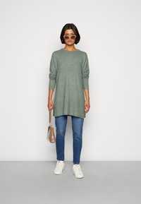 Suéter verde de malha com mangas longas, fendas laterais, combinado com jeans azuis e ténis brancos. A segurar uma mala transparente, usando óculos de sol.