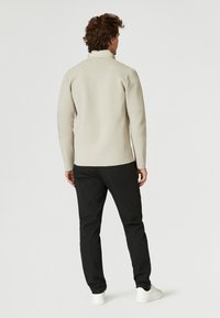 Beige stickad tröja med en delad hals, med en texturerad baksida och tonade axelaccenter, tillsammans med svarta byxor och vita sneakers.