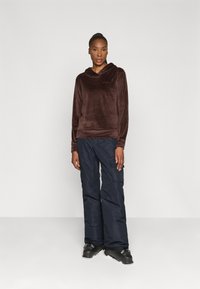 Brauner Velours-Hoodie mit Kapuze und langen Ärmeln, kombiniert mit dunkelblauen Cargo-Hosen mit seitlichen Taschen. Schwarze Schuhe runden das Outfit ab.
