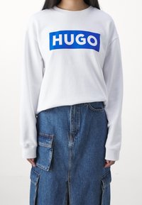 Felpa bianca a maniche lunghe con logo rettangolare blu "HUGO", abbinata a una gonna in denim con tasche laterali e uno spacco frontale.